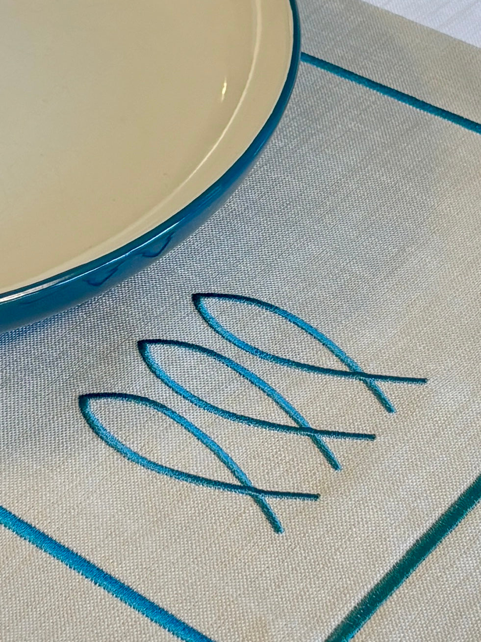 14x20 Inches35x50 Centimeters Trifin nautical table mat in Sand color, featuring three embroidered fish motifs on Sand color fabric with a white border. Designed for life at sea, it offers a balance of coastal sophistication and everyday practicality for onboard dining experiences.